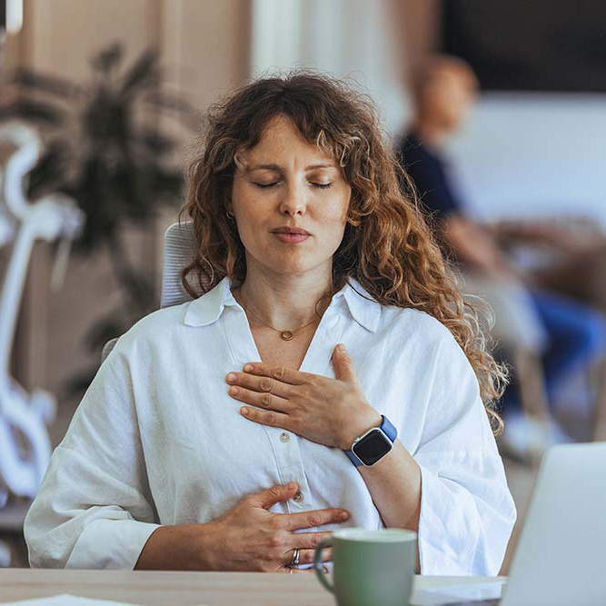 Vrouw in witte blouse aan tafel zittend met handen op borst en buik en ogen gesloten en op tafel staan een groene mok en laptop.