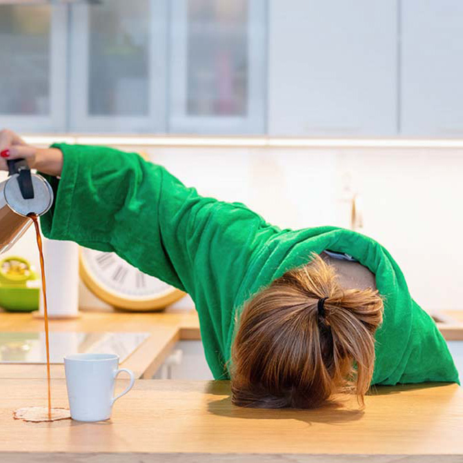 Vrouw met groene trui ligt met hoofd op een tafel en schenkt met hand koffie naast een kopje